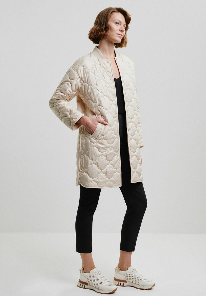 Beżowy pikowany płaszcz z kołnierzem stojącym, długimi rękawami i bocznymi kieszeniami, zestawiony z czarnymi spodniami i jasnymi trampkami z perforacjami.