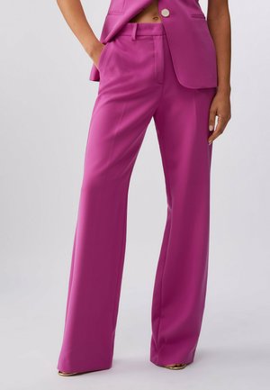 Donna che indossa pantaloni a vita alta e gamba larga color magenta con cuciture sartoriali, abbinati a un blazer dello stesso colore e tacchi aperti dorati.