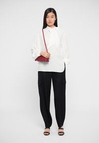 White blouse with a large bow tie collar, paired with black cargo pants featuring side pockets. Model holds a small burgundy crossbody bag.