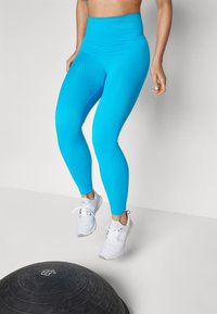 Blå ribbede leggings med høy midje, sammen med hvite treningssko, vist mot en lys bakgrunn. Treningsutstyr nedenfor.