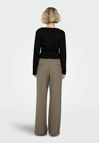 Ženska s kratkimi blond lasmi stoji s hrbtom proti gledalcu, oblečena v oprijeto črno majico z dolgimi rokavi in ohlapne olivno zelene hlače z zadnjimi žepi.