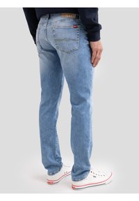 Férfi világoskék farmerral és fehér sportcipővel, piros és fekete részletekkel, világosszürke háttér előtt állva.