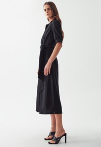 Vestito midi nero con maniche a sbuffo corte, vita con cintura e spacco laterale. Indossato con sandali neri con tacco alto, tessuto liscio.