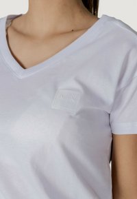 T-shirt bianco in cotone con scollatura a V, caratterizzato da un dettaglio logo in rilievo nell'angolo superiore sinistro. Texture morbida, design aderente.