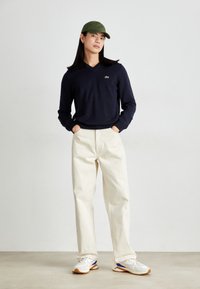 Námornícky svetrier s V-výstrihom, krémové široké nohavice, zelená čiapka a biele tenisky s modro-oranžovými akcentmi. Hladká textura, voľný strih.