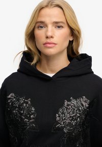 Sudadera negra con grandes diseños florales adornados con lentejuelas en la parte delantera. Tela suave con un bolsillo tipo canguro y capucha con cordón.