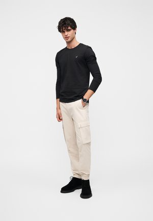 Schwarzes Langarmshirt, beige Cargohosen mit Seitentaschen und schwarze Wildlederstiefel. Das Modell steht vor einem schlichten weißen Hintergrund.
