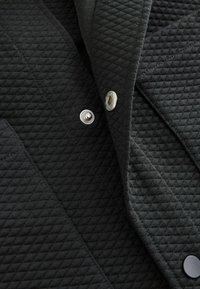 Černá prošívaná látka s diamantovým vzorem, vybavená stříbrnými patentními knoflíky a hladkou texturou. Design zahrnuje překrývající se vrstvy.