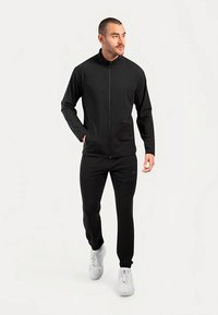 Veste noire zippée avec un col montant, assortie à un pantalon noir. Les deux vêtements sont confectionnés dans un matériau lisse et léger.