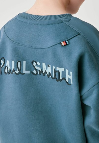 Sinine sviiter, mille seljal on kontrastsetes värvides "Paul Smith" logo, ribatexdikuga krae ja õlal mitmevärviline kangast silt.