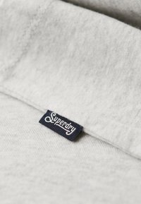 Jasnoszara dzianina dresowa o miękkiej, teksturowanej powierzchni. Posiada granatową metkę z napisem "Superdry" w białej, kursywnej czcionce.