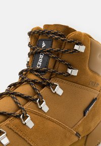 Brown suede and textile hiking boot with black and orange laces threaded through metal eyelets and a black logo label on the tongue.