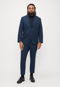 Marineblauw pak met een enkelsluitend jasje met twee voorzakken en bijpassende slim-fit broek, gecombineerd met een zwart overhemd en schoenen.