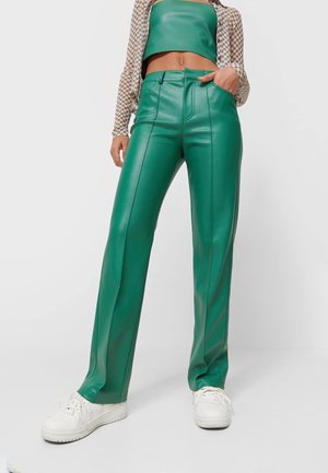 Personne portant un pantalon en cuir vert taille haute, un haut vert sans bretelles, un shrug à manches longues à carreaux et des baskets blanches, debout avec une main dans la poche.
