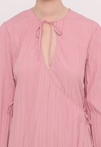 Robe plissée rose avec un col en V et une fermeture à nouer au niveau du col, dotée de manches longues et de liens latéraux pour un ajustement réglable.