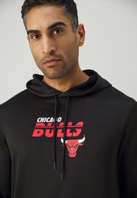 Hoodie noir avec cordon de serrage, présentant un graphisme et un logo rouge "CHICAGO BULLS". Tissu lisse, design moderne et impression audacieuse.