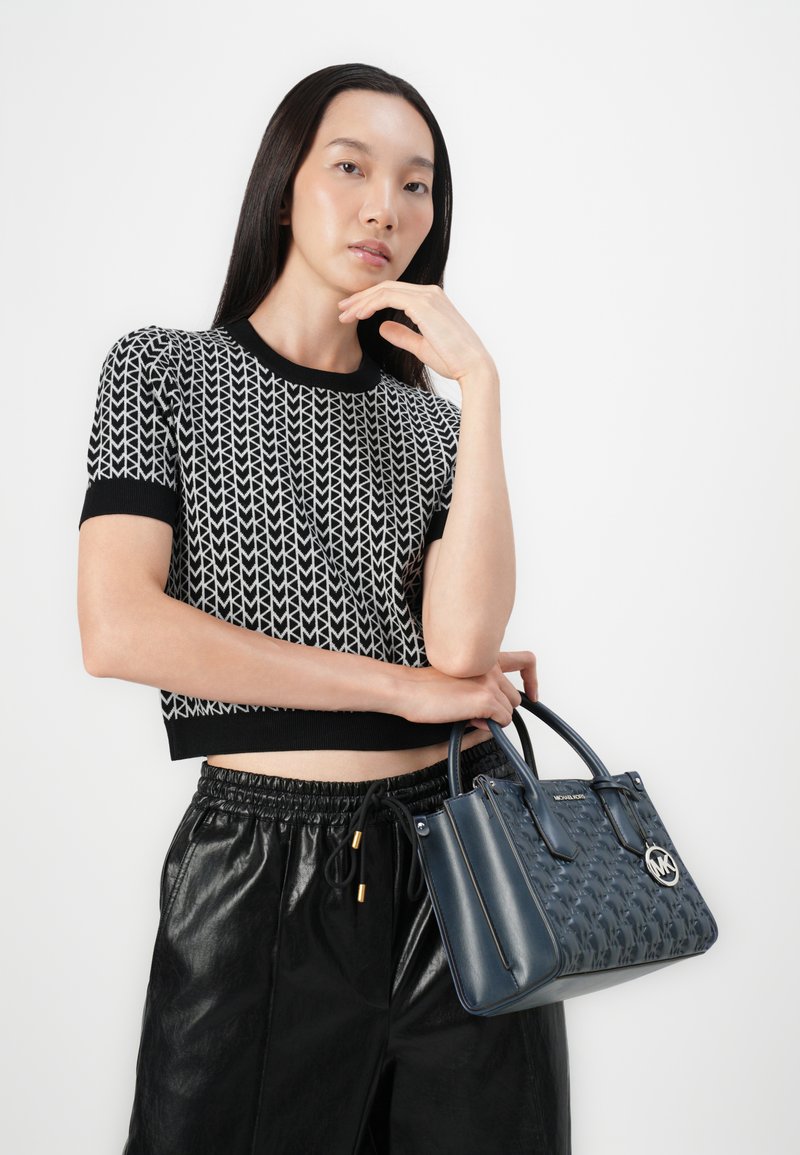 Borsa a tracolla in pelle blu con design intrecciato, maniglie doppie e logo argentato. Il modello indossa un crop top a righe bianche e nere e pantaloni neri.