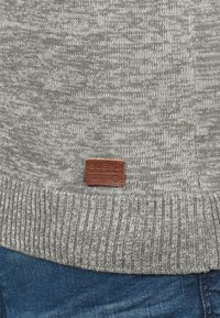 Szary dzianinowy sweter z prążkowanym wykończeniem i małą brązową naszywką z logo na dole, na tle niebieskiego denimu.