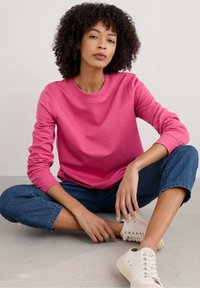 Vrouw met krullend haar, gekleed in een roze shirt met lange mouwen, blauwe jeans en witte sneakers, zittend met gekruiste benen op een grijze vloer tegen een effen achtergrond.