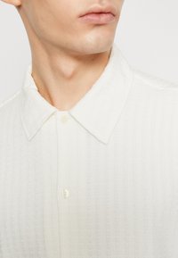 Vit texturerad skjorta med krage, med synliga sömmar och knäppning, tillverkad av lättviktsmaterial.