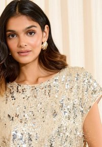 Kultainen paljettitoppi, jossa on hihat, ominaisuutena kimalteleva tekstuuri ja väljä istuvuus. Huomaamaton pystysuuntainen paljettikuosis tuo syvyyttä.