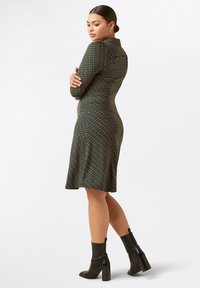 Vestido verde con estampado que presenta una silueta ajustada, mangas largas, cuello y un dobladillo de longitud media. Combinado con botas de tacón negras.