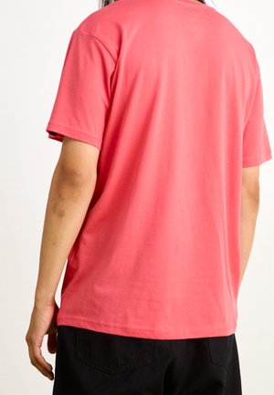 Persoon met een effen koraalkleurig T-shirt met korte mouwen en een zwarte broek, van achteren gezien tegen een witte achtergrond.