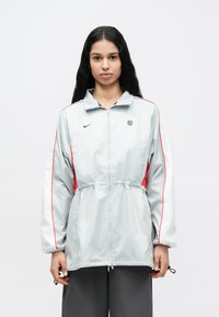 Helehall tikandiga lukuga jakk, millel punased küljeservad ja logo. Vööjoon on nööriga reguleeritav ning varrukad on pikad ja veidi padjastatud. Pind on sile.