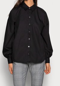 Black button-up shirt with a classic collar, long puffed sleeves, and front button closure, paired with gray plaid patterned trousers.