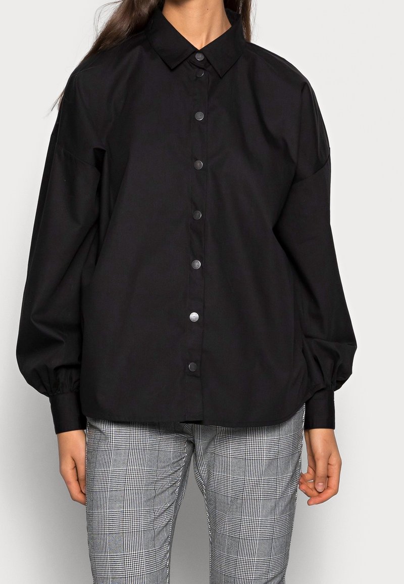 Black button-up shirt with a classic collar, long puffed sleeves, and front button closure, paired with gray plaid patterned trousers.