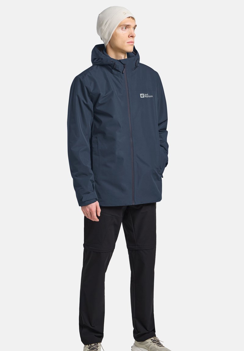 Giacca impermeabile della marina con cappuccio, zip frontale e logo. Abbinata a pantaloni neri con tasche e tessuto testurizzato. Berretto neutro indossato.