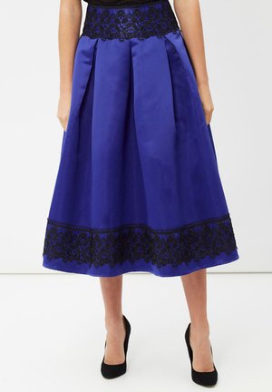 Mujer con falda midi azul con encaje negro en la cintura y el dobladillo, combinada con tacones altos negros, de pie sobre un suelo blanco.