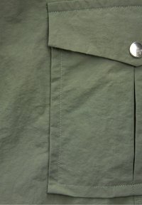 Nahaufnahme einer olivgrünen Stofftasche mit Klappe, die mit einem einzigen silbernen Druckknopf verschlossen ist, sowie sichtbaren Nähten entlang der Kanten.