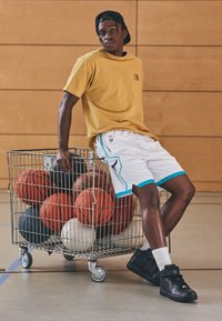 Yellow short-sleeve shirt, white athletic shorts with blue accents, black sneakers. A wire cart holds various basketballs in different colors.