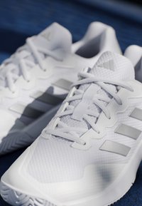 White athletic shoes with laces and gray three-stripe logo on the sides, placed on a dark blue textured surface.