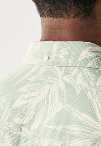 Camicia a maniche corte color verde chiaro, realizzata in tessuto strutturato con motivo di foglie tropicali bianche. Presenta un colletto con bottoni e un dettaglio sulla schiena.