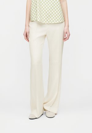 Žena v krémových širokých nohaviciach s vysokým pásom, v kombinácii s béžovými topánkami a svetlozeleným vzorovaným topom.