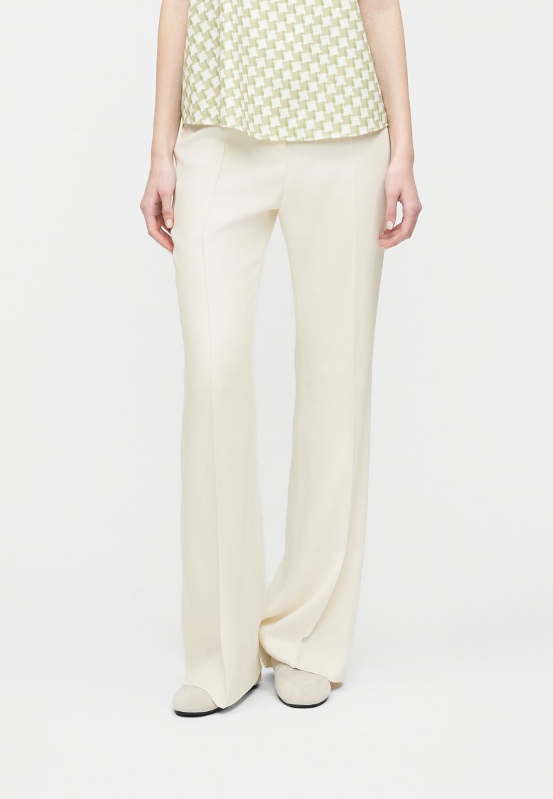 Γυναίκα που φοράει μπεζ παντελόνι με ευρύ ποδόγυρο, ψηλόμεσο, συνδυασμένο με μπεζ παπούτσια και ανοιχτοπράσινο τοπ με σχέδια.