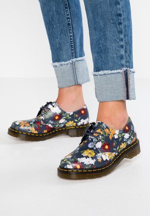Marineblå blomstrede snøresko med gul syning, båret med opkrogede blå jeans på en hvid overflade.