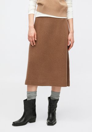 Personne portant une jupe midi en tricot marron, un pull sans manches beige sur un haut blanc à manches longues, des chaussettes grises et des bottes en cuir noires mi-mollet.
