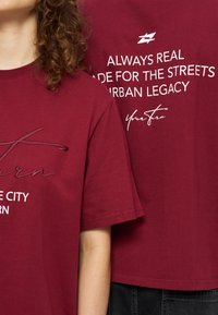 Δύο άτομα φοράνε κόκκινα T-shirts με λευκό κείμενο για την αστική κληρονομιά και τους δρόμους, ο ένας δείχνει το μπροστινό μέρος, ο άλλος το σχέδιο στο πίσω μέρος.