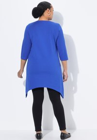 Blaue Tunika mit Dreiviertelärmeln, mit einem abgerundeten Saum, kombiniert mit schwarzen Leggings und Ballerinas. Glatte Stoffoberfläche.