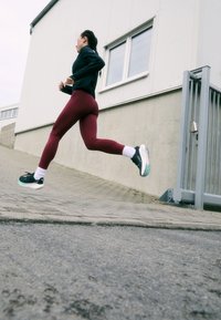 Mujer con leggins burdeos y top negro corriendo colina arriba por una acera pavimentada junto a un edificio blanco con ventana y reja de metal.