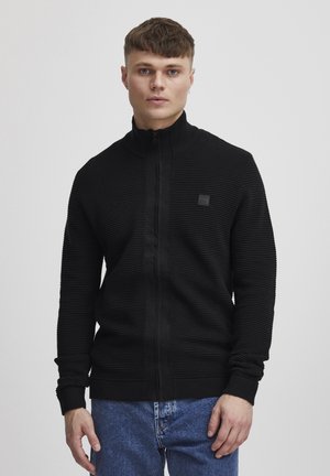 Sort zip-up cardigan med ribbet struktur og standard krave. Indeholder et lille logo-patch på brystet og tætsiddende manchetter.