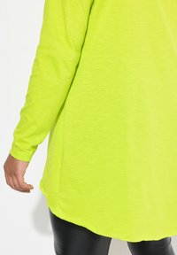 Langärmlige neongrüne Tunika mit abgerundetem Saum, aus weichem Material, kombiniert mit schwarzen Leggings.