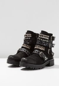 Botas negras hasta el tobillo hechas de ante y cuero. Presentan múltiples correas con tachuelas y hebillas, detalles texturizados y una suela de tracción gruesa.