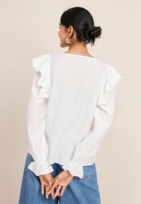 Blusa bianca con spalle arricciate e maniche lunghe, con bordi in pizzo, abbinata a jeans in denim blu. Il tessuto sembra leggero e dalla texture interessante.