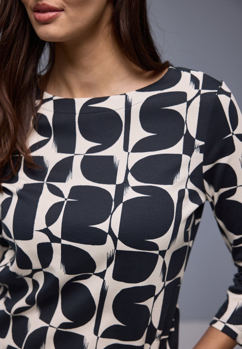 Femme portant un haut à motifs géométriques noirs et blancs, avec un col rond et des manches trois-quarts, sur un fond gris.