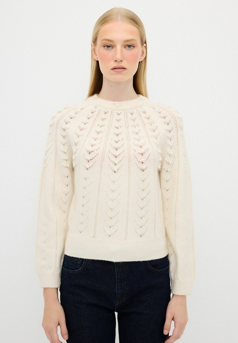 Maglione lavorato a maglia crema con un motivo testurizzato di frecce e piccole perle, caratterizzato da un collo rotondo e maniche lunghe. Abbinato a jeans scuri.