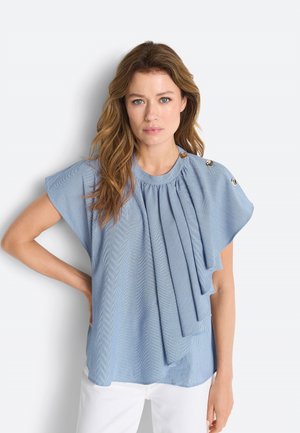 Naine laineliste juustega, kes kannab helesinist voltidega toppi kuldsete nööpidega õlal ja valgeid pükse, seisab valge tausta ees.